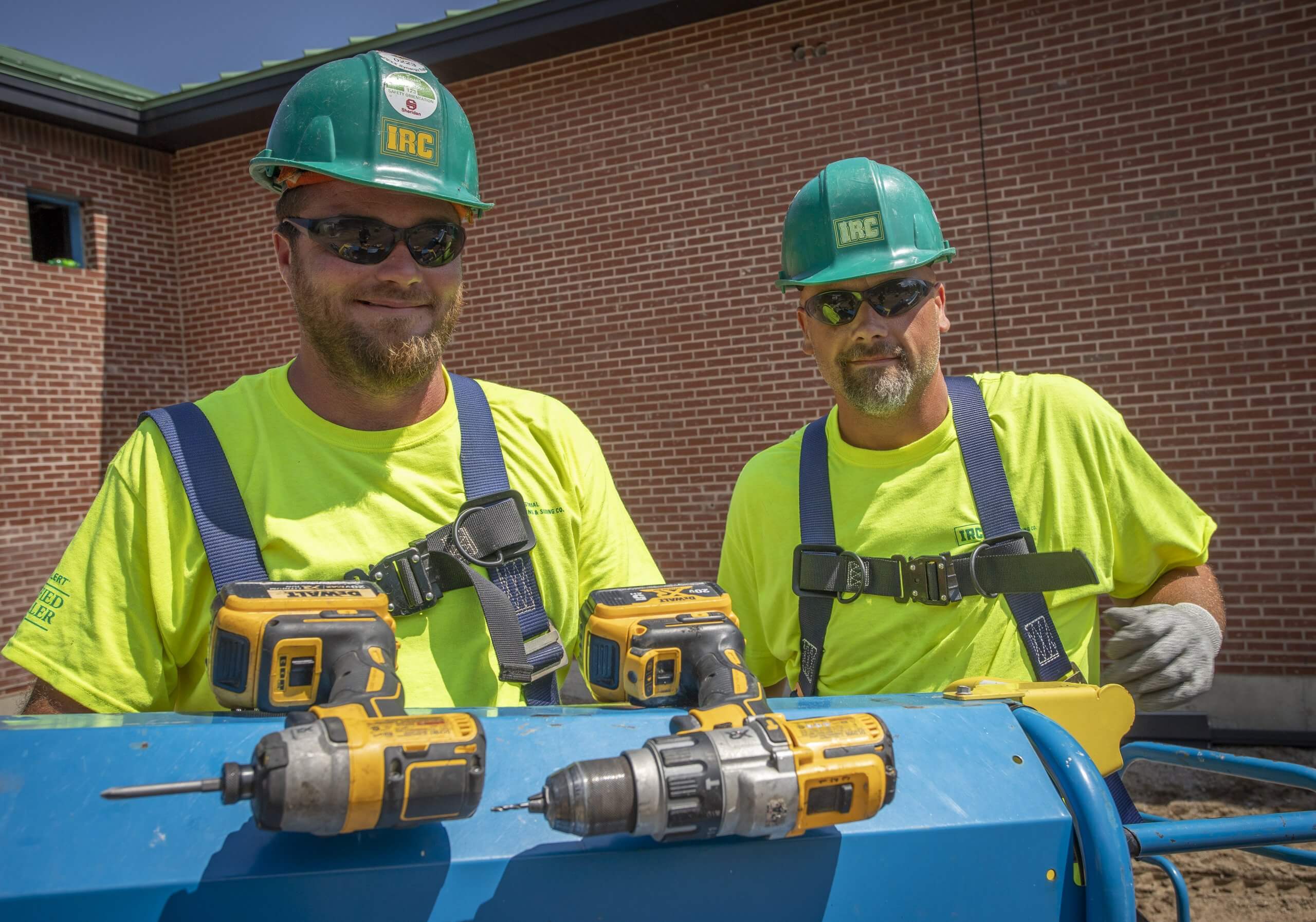 Two roofing technicians smile while at a job site wearing hardhats, safety harnesses and with tools